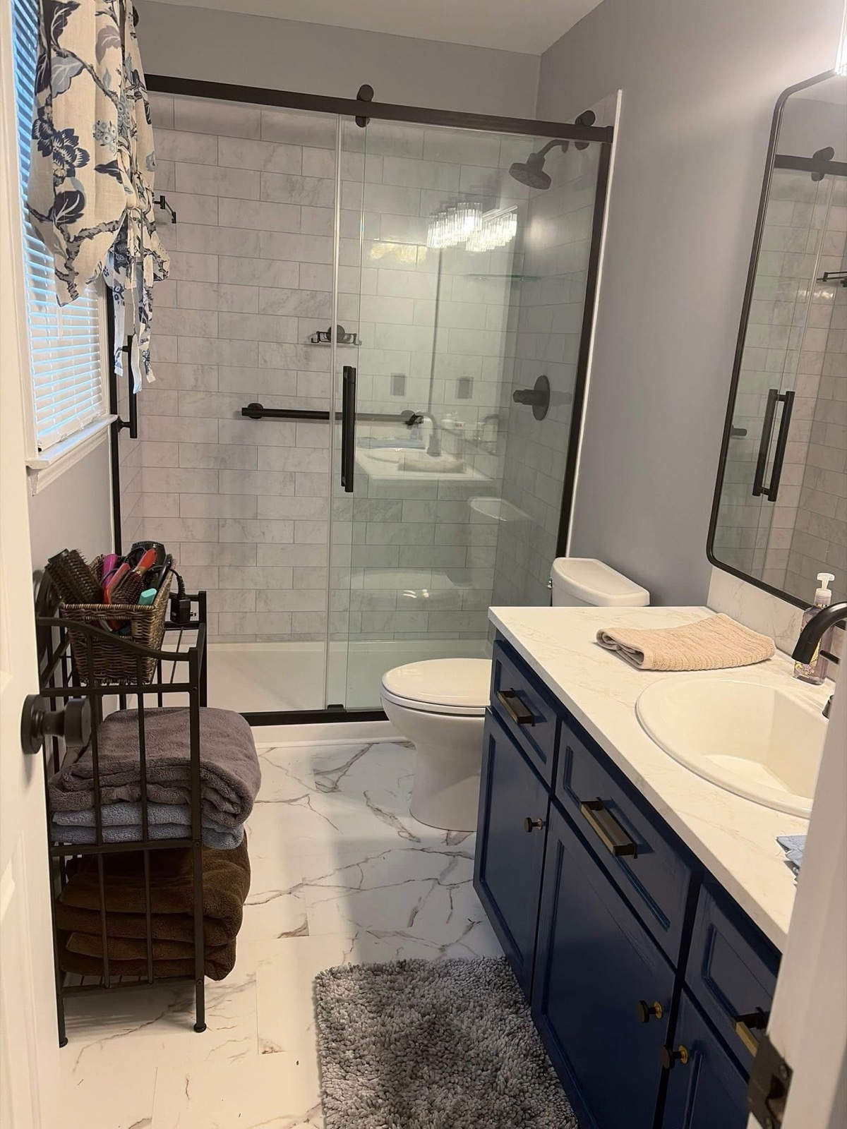 The same bathroom after remodeling with a glass shower enclosure, blue vanity, and marble-look tile floor.